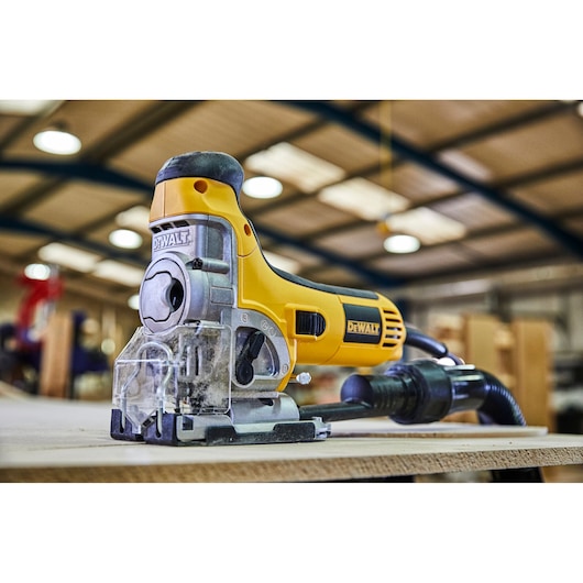 DEWALT electric jigsaw power tool in yellow and black, positioned on a wooden surface in a workshop setting. The tool is plugged in and appears ready for use.