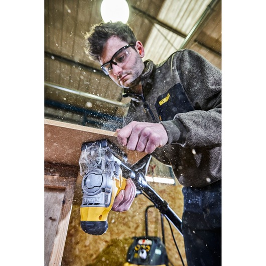 Person using a DEWALT jigsaw to cut a wooden board in a workshop. Sawdust is flying in the air, and the person is wearing a DEWALT jacket.