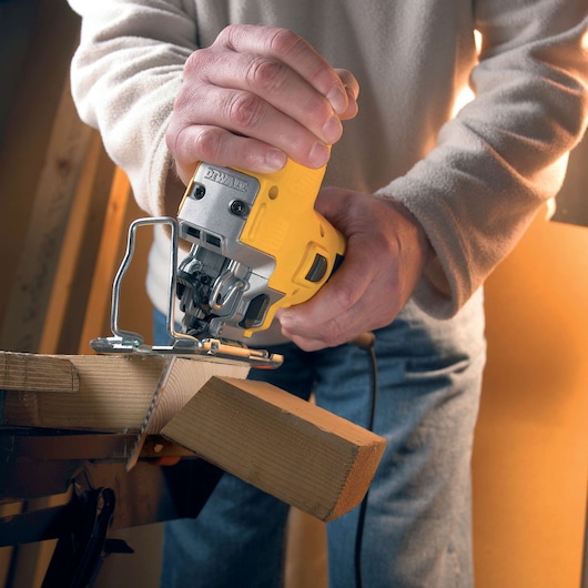 A person is using a yellow DEWALT jigsaw to cut a piece of wood. The jigsaw is powered and the user is holding the tool