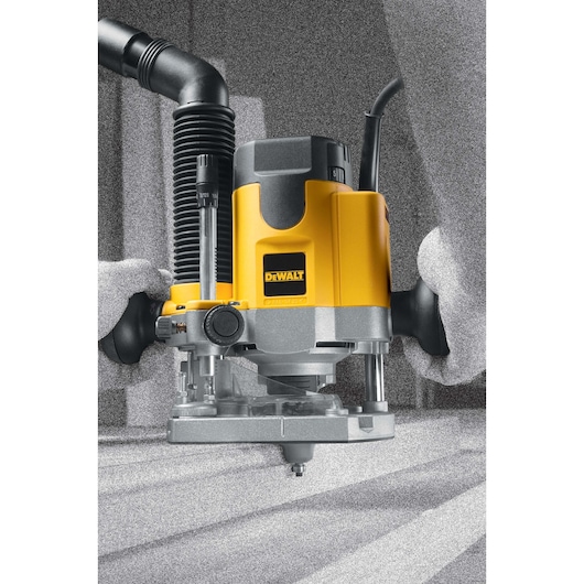 A DEWALT DW621 plunge router is being operated, with a yellow and black body, attached to a hose for dust extraction. The router is being used to work on a wooden surface.