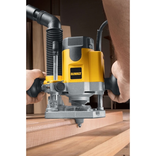 Close-up of a DEWALT DW621 router being used on a wooden surface, with hands holding the tool and a dust extraction hose attached.