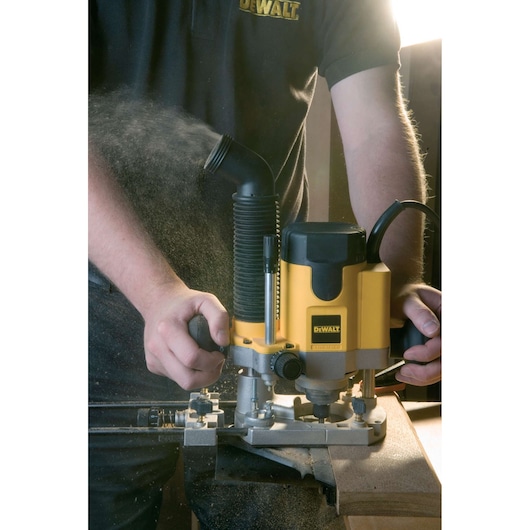 A person using a DEWALT DW621 router tool to work on a piece of wood, with wood dust being expelled through a dust extraction port. The router is yellow and black, and the operator is wearing a DEWALT-branded shirt.