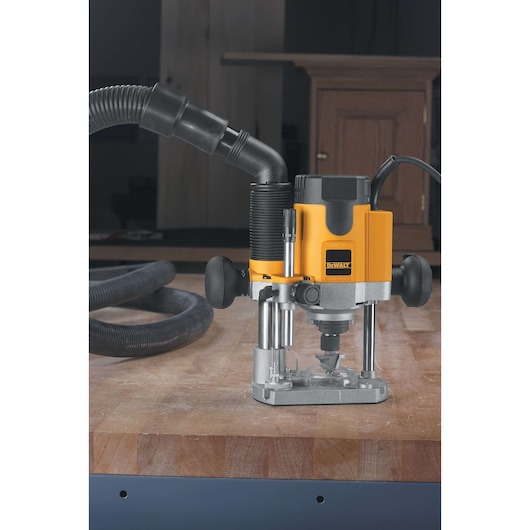 A DEWALT plunge router is positioned on a wooden workbench. The router has a yellow and black body, a dust extraction hose attached, and two black handles. The workspace is indoors with woodworking materials visible in the background.