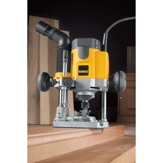A DEWALT plunge router tool, model DW621, is shown placed on a wooden surface. The tool features a yellow and black casing, two side handles, and a dust extraction attachment. It is designed for precision woodworking and carpentry tasks.