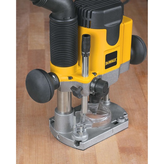 A DEWALT plunge router with a yellow and black body placed on a wooden surface. The tool features ergonomic handles, adjustment knobs, and a dust extraction port for woodworking tasks.