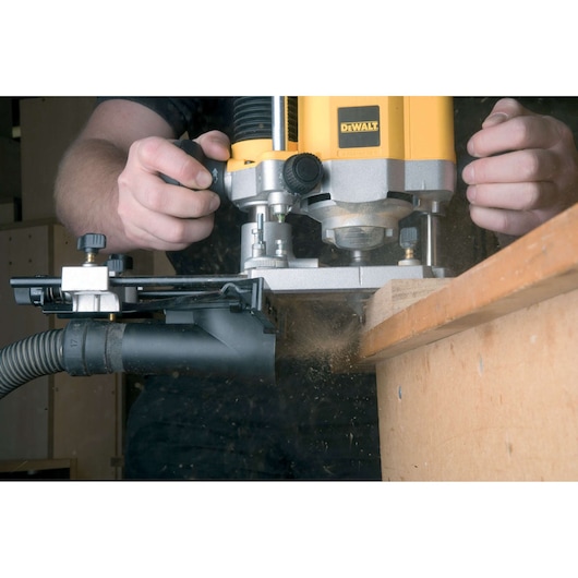 A person using a DEWALT plunge router to shape the edge of a wooden board, with sawdust being collected by an attached dust extraction system.