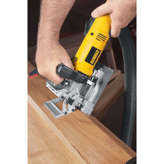 A person using a DEWALT DW682K biscuit joiner to cut a slot into the edge of a wooden board. The tool has a yellow and black body with adjustment dials visible, and is connected to a vacuum hose for dust extraction.