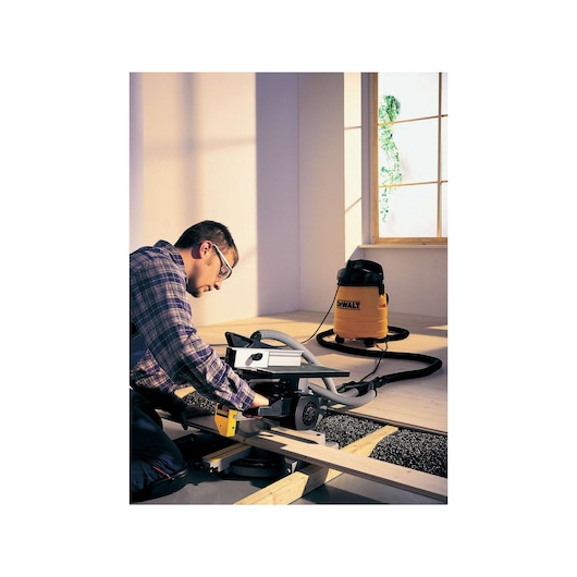 A person using a DEWALT DW711 power tool to cut wood planks indoors. There is a yellow DEWALT vacuum connected via hose in the background, next to a window.