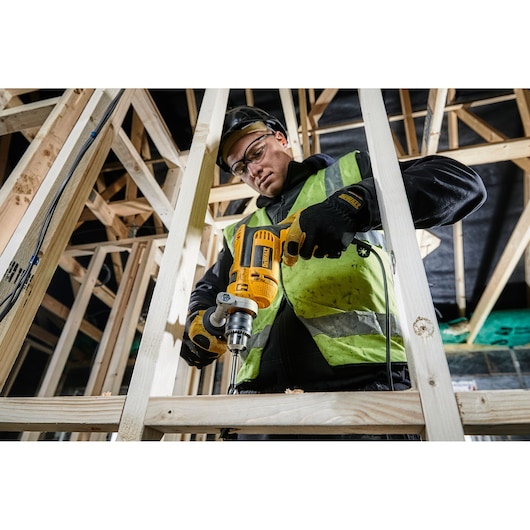 A person wearing a safety vest and gloves is using a yellow DEWALT drill (model DWD024K) to drill into a wooden frame at a construction site.