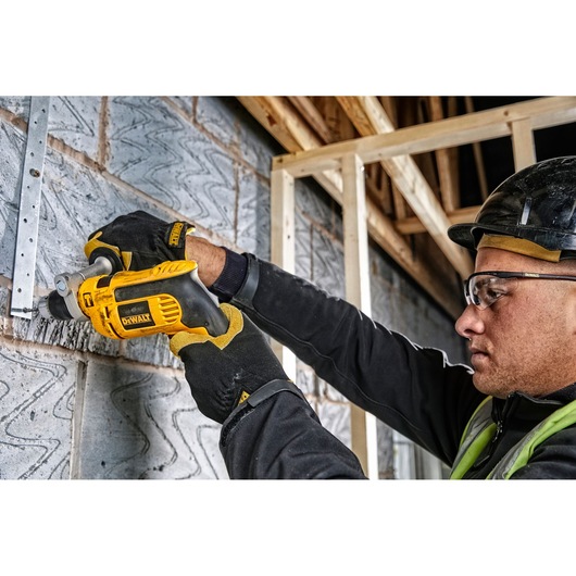 A person wearing construction gloves uses a yellow DEWALT power drill to drill into a brick wall at a construction site. The background shows unfinished wooden framing and a partially built structure. The person's face is blurred for privacy.