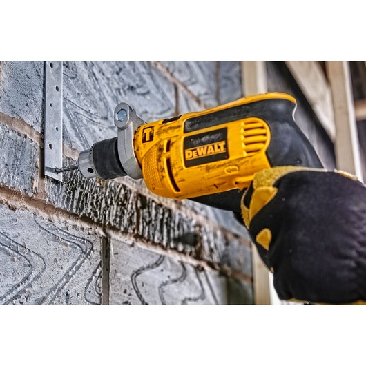 A DEWALT electric drill is being used to drill a hole into a metal bracket attached to a cement block wall. The drill is held by a gloved hand.