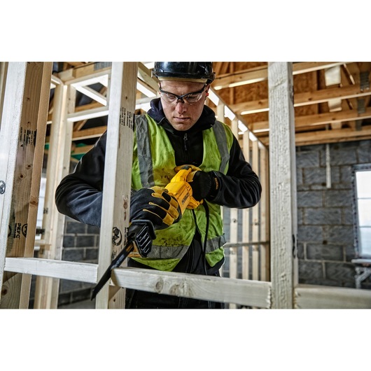 A construction worker wearing a helmet, gloves, and a high-visibility vest uses a yellow DEWALT reciprocating saw (DWE305PK) to cut wooden beams inside a building under construction. The worker's face is blurred for privacy.