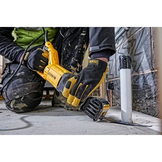 A worker wearing black and yellow DEWALT gloves uses a DEWALT DWE305PK reciprocating saw to cut a metal pipe close to the floor in an indoor construction setting.