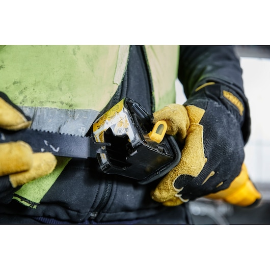 Close-up image of a person wearing protective gloves and a yellow-green safety vest, holding a DEWALT reciprocating saw with a blade. The saw is being positioned near the waist, showing work preparation.