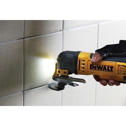 A person uses a yellow and black DEWALT oscillating multi-tool to cut or remove grout from between white wall tiles. The