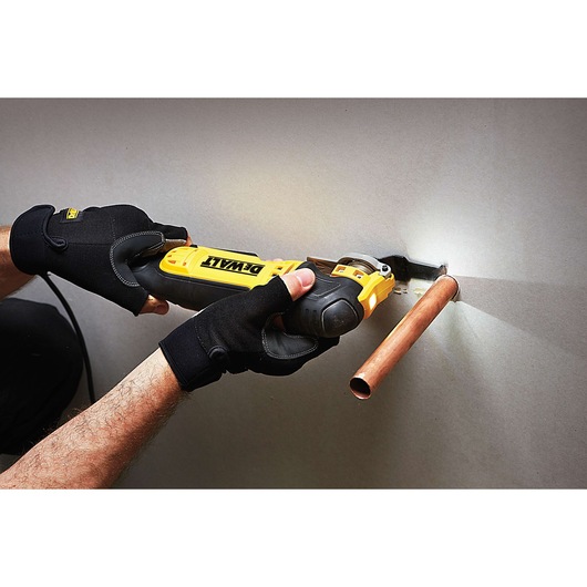 A person wearing black gloves uses a DEWALT multi-tool to cut drywall around a copper pipe.