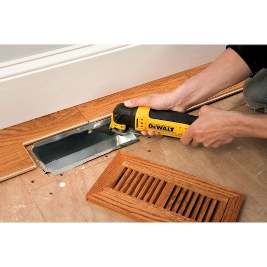 A person is using a DEWALT oscillating multi-tool to cut wood flooring around a metal floor vent opening. The wooden vent