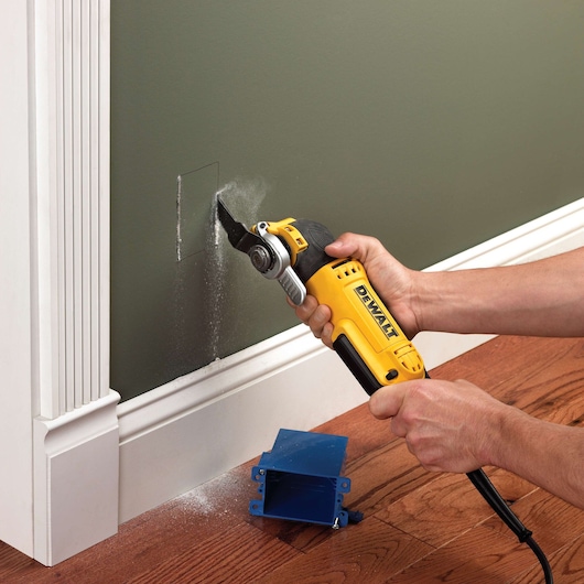 A person uses a yellow DEWALT DWE315KT multi-tool to cut a rectangular hole in a wall near the baseboard. Dust is visible as