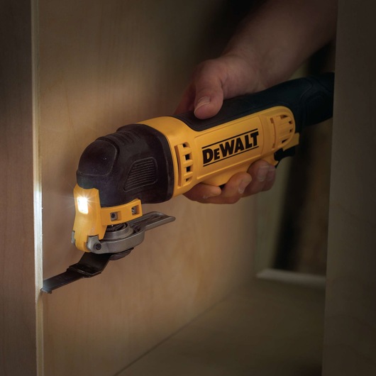 A DEWALT oscillating multi-tool with a yellow and black design is being used to cut wood. A built-in LED light illuminates