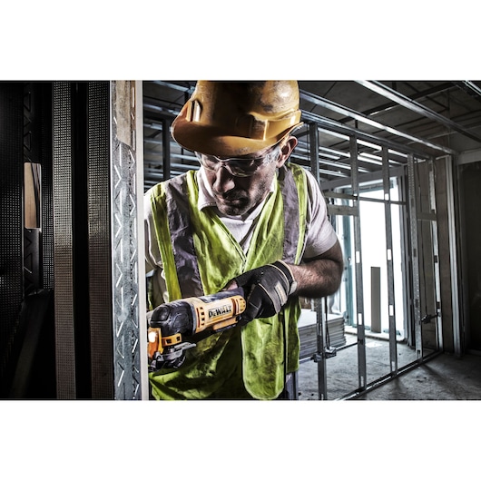 A construction worker wearing a yellow hard hat and a high-visibility vest is using a DEWALT DWE315 oscillating multi-tool to cut metal framing at a building site. The worker's face is blurred for privacy.