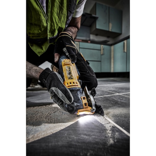 A person wearing work gloves uses a DEWALT DWE315 multi-tool to cut or grind material between floor tiles, with a bright LED light illuminating the work area.