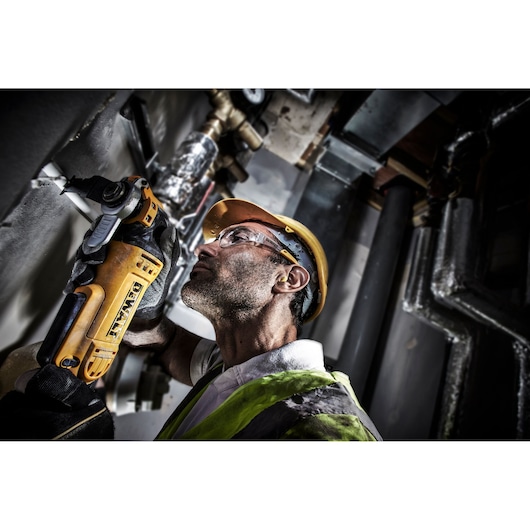A person wearing a yellow hard hat and safety vest uses a DEWALT DWE315 power tool to cut or grind material on a wall in an industrial setting. Their face is obscured.