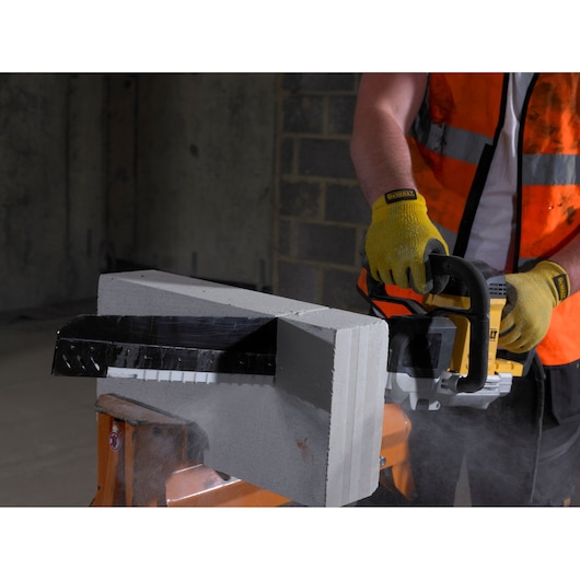 A person wearing yellow gloves and an orange safety vest is using a DEWALT power saw to cut through a large concrete block