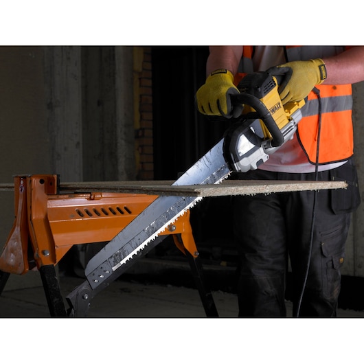 A person wearing yellow gloves and an orange safety vest is using a DEWALT electric saw to cut a piece of wood placed on an orange workbench.