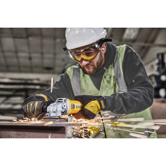 A worker wearing a safety helmet and gloves is using a DEWALT DWE4036 angle grinder to cut metal, with sparks flying.