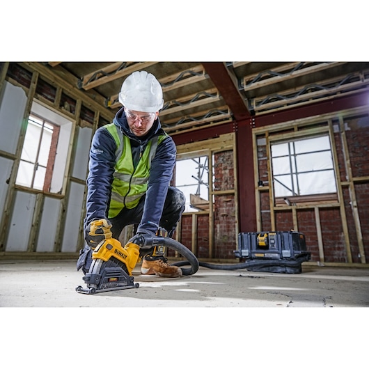 A construction worker wearing a high-visibility vest and hard hat is using a DeWalt power tool on the floor of a partially built room. The worker is kneeling, and the tool is connected to a hose. There is a DeWalt equipment case in the background.