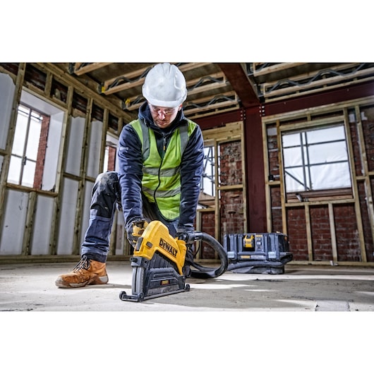 A person wearing a safety helmet, high-visibility vest, and work boots uses a DEWALT DWE46229 power tool on a construction