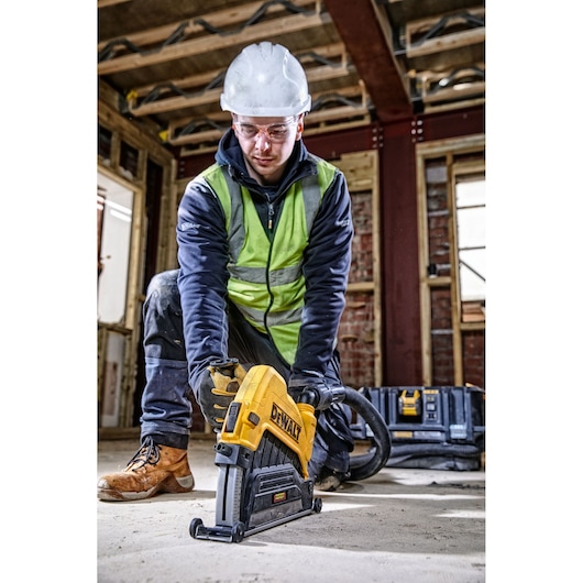 A construction worker wearing a high-visibility vest, helmet, and gloves is kneeling in an unfinished building using a