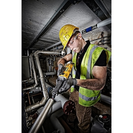 A worker wearing a yellow hard hat, safety vest, and gloves is using a DEWALT DWE4884 power tool to grind or polish a metal