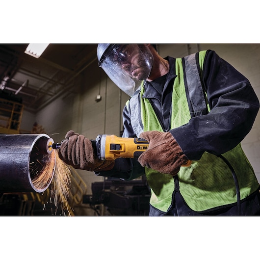 A person wearing safety gear uses a DEWALT power tool to grind or cut a metal pipe, producing sparks.