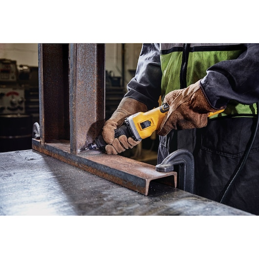Close-up of a person wearing gloves and a safety vest using a yellow DEWALT power tool to grind or polish a metal beam in an industrial workshop.