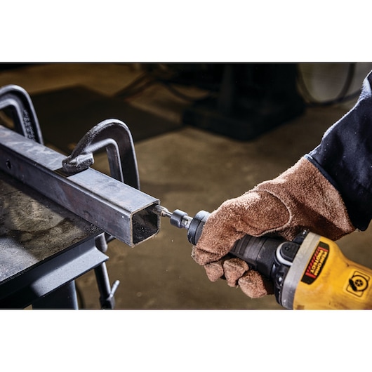 A person wearing brown protective gloves is using a yellow DEWALT power tool to grind or smooth the edge of a metal rectangular tube that is clamped to a workbench.