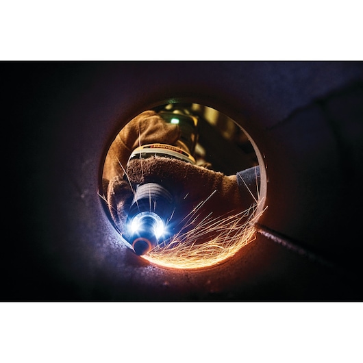 A close-up view inside a metal pipe showing a gloved hand using a DEWALT power tool to grind the inner surface, creating bright sparks.
