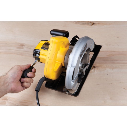 A person uses a screwdriver to adjust a yellow DEWALT circular saw placed on a wooden surface.