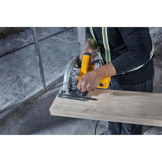 A person using a yellow DEWALT circular saw (model DWE5615) to cut a wooden plank, with sawdust and safety gear visible.