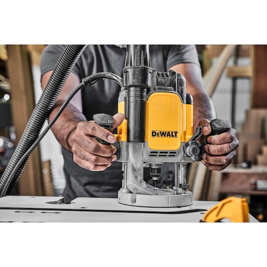 A close-up of a person operating a yellow DEWALT plunge router tool on a workbench in a workshop. The person's hands are