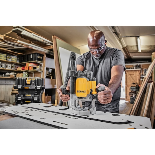 A person using a DEWALT DWE625KT plunge router on a workbench in a woodworking workshop. The router is yellow and black, and
