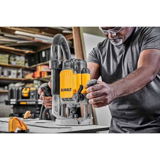 A person is operating a DEWALT DWE625KT plunge router on a workbench in a workshop. The router is yellow and black and is