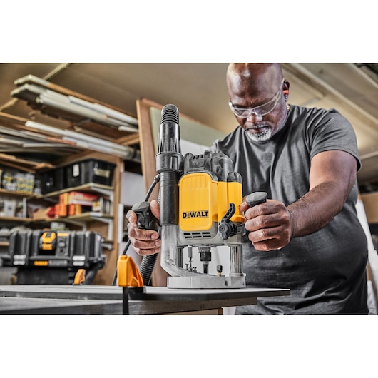 A person using a DEWALT DWE625KT plunge router on a worktable in a workshop, with the tool's yellow and black design clearly
