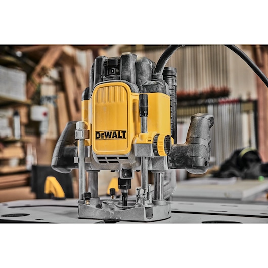 A DEWALT DWE625 plunge router is placed on a workbench in a woodworking workshop. The router has prominent yellow and black coloring, ergonomic handles, and adjustment knobs. Various woodworking tools and materials are visible in the background.
