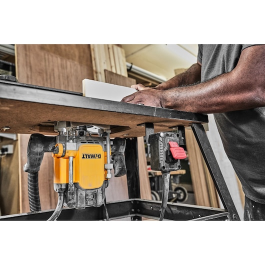 A DEWALT router mounted under a tabletop in a workshop, with a person guiding a piece of wood along the table for precision