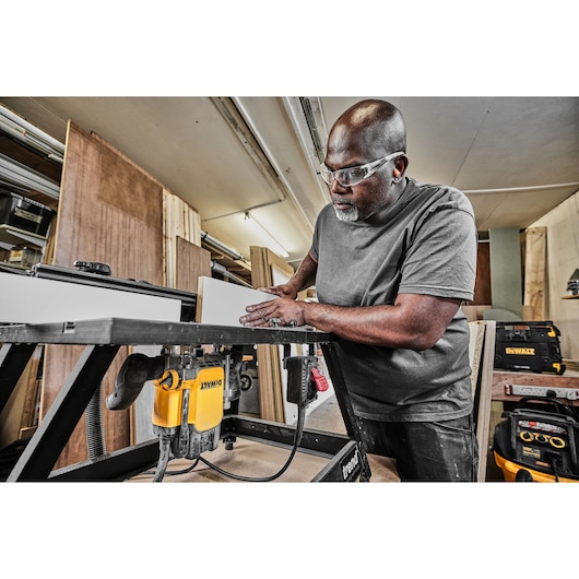 A person working in a woodworking shop using a DEWALT DWE627KT router mounted on a workbench. Various DEWALT tool cases and