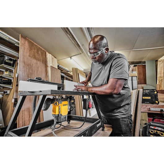 A person working in a woodworking workshop using a DEWALT DWE627KT router mounted on a workbench, surrounded by wood panels