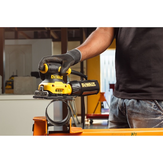 Close-up of hands connecting a dust extraction hose to a yellow DEWALT DWE6411 power sander in a workshop setting.