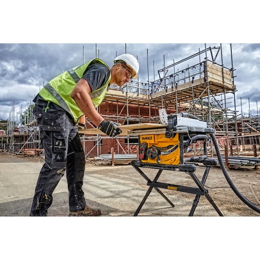 A construction worker wearing a safety vest and helmet uses a DEWALT DWE7485WS table saw on a stand at a building site with scaffolding and materials in the background.