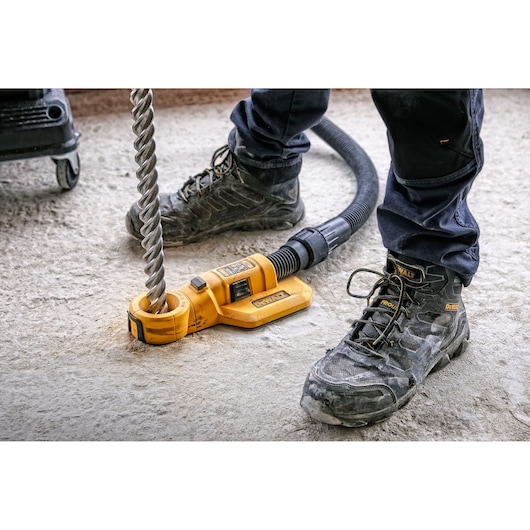 A DEWALT DWH050 dust extraction attachment is connected to a vacuum hose and positioned around a large drill bit on a construction site floor. The feet of a person wearing DEWALT branded work boots and dark pants are visible.